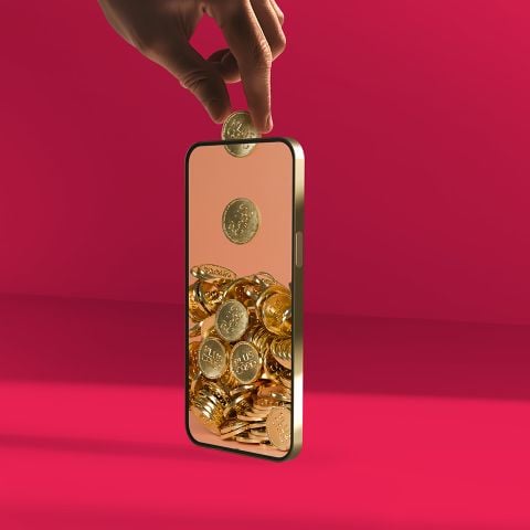 Eine Hand lässt eine Goldmünze in ein Smartphone fallen, in dem sich bereits ein Haufen Goldmünzen ansammelt.