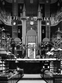 Eine Schwarz-Weiß-Aufnahme des Innenraums eines Kaufhauses, auf der eine zentrale Ausstellungsfläche mit zwei hohen, spitzen Türmen zu sehen ist, über der das Wort „TEPPICHE“ hängt.